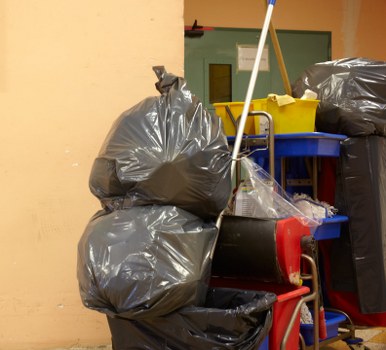 The image displays an indoor area with a concrete or tile floor, containing multiple large black plastic bags filled with rubbish, stacked against a light-colored wall. Two of the bags are positioned at the forefront, with one resting on a red trolley or cart. To the right, there is a blue shelving unit holding a yellow plastic bin or container and a small white bucket, with a mop or broom leaning against it. A partially visible door with a green frame is in the background, behind the shelving. The setting suggests a space used for waste collection or storage, consistent with rubbish removal services in the Dalston area. The environment appears well-organized for rubbish disposal, with an emphasis on functional storage and waste segregation. The lighting is even, illuminating the surfaces clearly and emphasizing the textures of the plastic bags, which are shiny and crinkled, and the various plastic containers. Commercial Waste Dalston's rubbish removal service could efficiently manage waste in this type of setting, typical for small storage or back-of-premises locations in London.