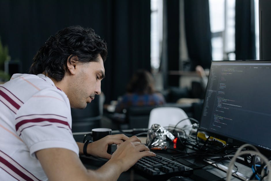 A young man with dark, wavy hair and light skin is seated at a desk indoors, concentrating on typing on a black keyboard. He is wearing a white shirt with maroon and gray horizontal stripes, which has